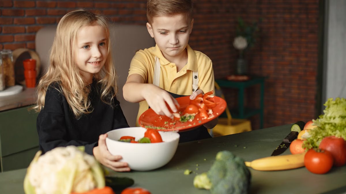 Positive little girl and boy mixing cutted ingredients for salad in big bowl while cooking at table with assorted fresh vegetables and fruits